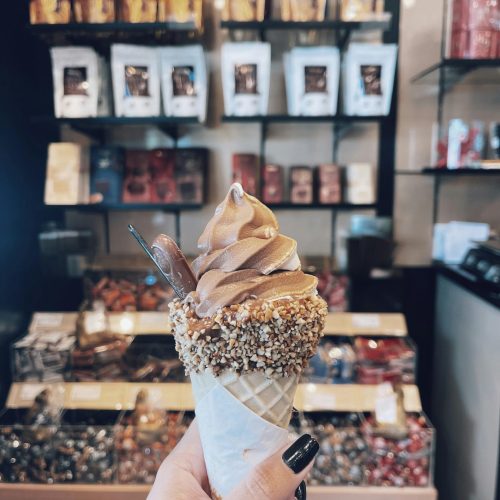 Close-up of a chocolate and vanilla swirl ice cream cone with nuts held in front of a candy store.
